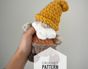 Puede incluir: Patrón de crochet para un pequeño gnomo con un cuerpo marrón, naranja y blanco y un gorro amarillo. El gnomo está siendo sostenido en una mano.