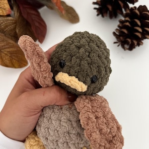 May include: A crocheted plush toy shaped like a bird with brown, green and beige fur. The bird has a small, yellow beak and black eyes.