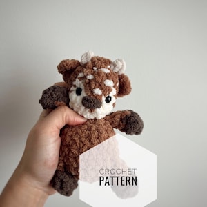 Puede incluir: Patrón de crochet para una vaca amigurumi con manchas marrones y blancas, ojos negros y nariz blanca. La vaca está siendo sostenida en una mano. El texto "CROCHET PATTERN" está en un hexágono blanco.