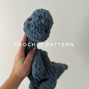 Puede incluir: Un juguete de dinosaurio azul de crochet con ojos negros. El juguete está siendo sostenido por una mano contra un fondo blanco. El texto "CROCHET PATTERN" es visible debajo del juguete.
