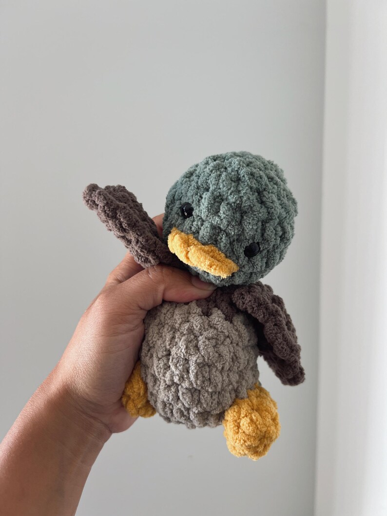 May include: A crocheted plush toy duck with a green head, brown body, and yellow beak and feet.
