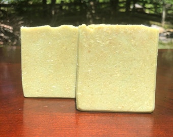 May include: Two bars of light green soap with a grainy texture. The soap bars are sitting on a wooden surface.