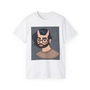 May include: A white short-sleeved t-shirt featuring a centered graphic illustration of a person with dark hair, blue highlights, small horns, round glasses, and a beard, wearing a brown shirt against a dark gray square background.
