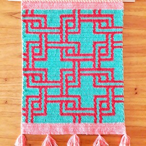 May include: A rectangular crochet wall hanging featuring a geometric maze pattern in bright red and turquoise. The piece is suspended from a thin wooden dowel and finished with a solid light pink border at the top and bottom, including five matching tassels.