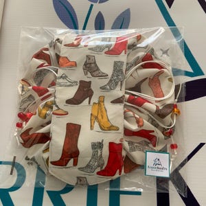 May include: A white drawstring bag featuring a pattern of different boots in red, brown, grey, and yellow. The bag is packaged in clear plastic and has a tag that reads "Karrie Krafts Handmade".