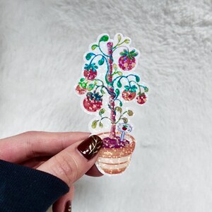 May include: A colorful sticker featuring a whimsical tree with red fruit and green leaves, planted in a decorative pot. The tree has a glittery effect, and a small sign with an apple is at the base of the tree.