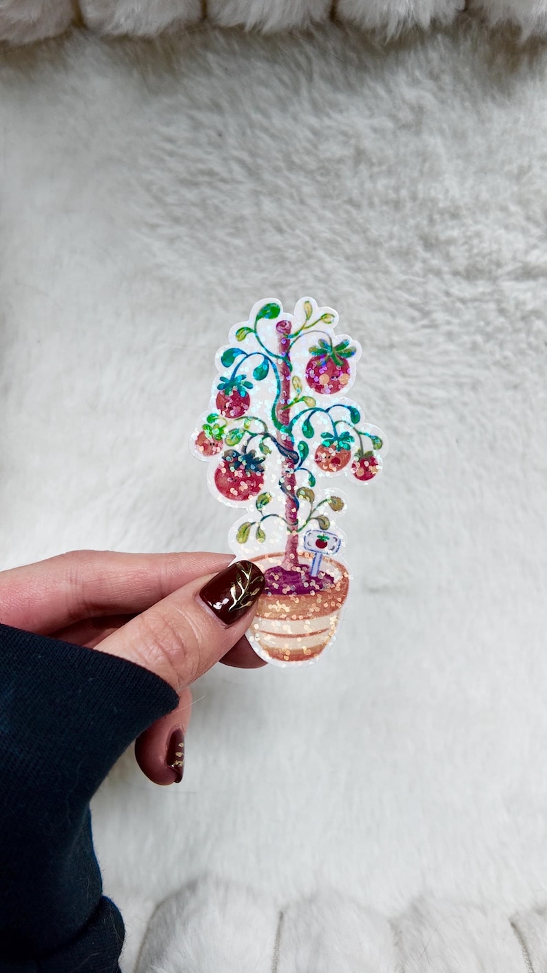 May include: A colorful sticker featuring a whimsical tree with red fruit and green leaves, planted in a decorative pot. The tree has a glittery effect, and a small sign with an apple is at the base of the tree.