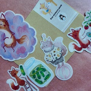 May include: A collection of whimsical fox stickers. The stickers feature various fox illustrations, including one in a cloud, another with a party hat, and one with a jar of pickles. The stickers are on a pink and tan background. The text "Fox stickers pack" is visible.