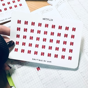 Peut inclure: Feuille d'autocollants avec le logo "N" rouge et le mot "Netflix" imprimé au-dessus. Les autocollants sont tenus au-dessus d'un agenda ouvert. Le texte "BOUTIQUE BY SHA" est imprimé.