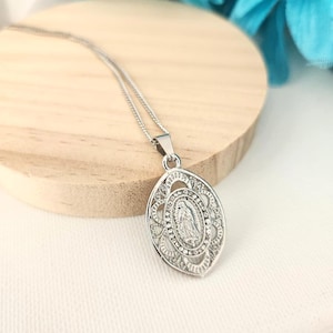 Puede incluir: Collar con colgante ovalado plateado con una figura religiosa detallada en un diseño de filigrana ornamentado. El colgante cuelga de una delicada cadena. El collar se exhibe sobre una superficie de madera.