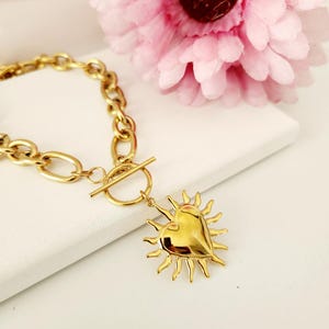Puede incluir: Un brazalete de cadena dorada con cierre de palanca y un colgante de corazón con rayos de sol.