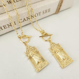 May include: Two gold necklaces with rectangular pendants depicting a religious figure. The pendants have detailed designs with a central figure and sunburst rays. The necklaces have a toggle clasp. The background is neutral.