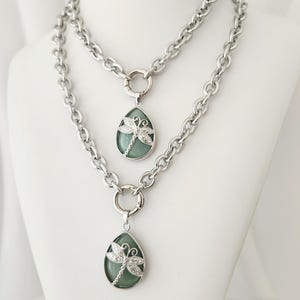 May include: Two silver-toned necklaces with a dragonfly pendant. Each necklace features a chunky chain and a teardrop-shaped pendant with a light green stone and a silver dragonfly design. The necklaces are displayed on a white mannequin.