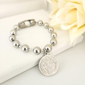 May include: A silver bracelet with a round charm featuring a religious image. The bracelet is made of small silver beads and has a lobster clasp.