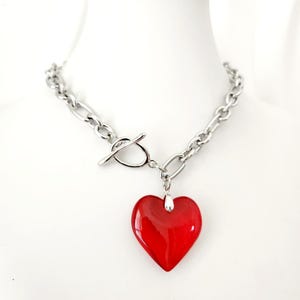 May include: A silver-toned chain necklace with a toggle clasp and a large, glossy red heart pendant. The heart is smooth and vibrant, suspended from a small silver bail. The necklace is displayed on a white surface.