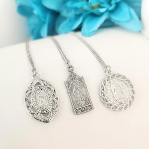 Puede incluir: Tres collares con colgantes plateados. Los colgantes presentan imágenes religiosas, incluyendo un diseño ovalado con una figura central, un colgante rectangular con una parte superior arqueada y un colgante redondo con un borde detallado. Sobre una superficie blanca.