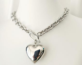 May include: A silver chain necklace with a large, round, silver heart pendant. The chain is a thick, curb style chain.