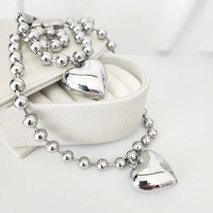 Puede incluir: Un collar de cadena de plata con dos colgantes en forma de corazón. El collar está hecho de pequeñas cuentas redondas y se muestra en una caja blanca.