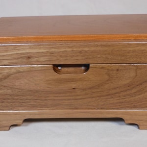 May include: A wooden box with a rectangular shape and a light brown finish. The box has a darker brown lid and a recessed handle on the front. The box sits on four small feet.