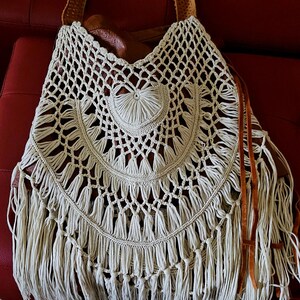 May include: Cream-colored crochet tote bag with a fringed bottom and a brown leather strap. The bag features a circular design with a heart-shaped detail in the center. The bag is made of a woven material.