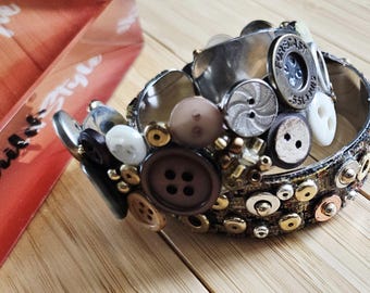 May include: A unique bracelet featuring a variety of buttons in shades of brown, white, and silver, accented with small gold-colored beads. The bracelet is set against a wooden surface.