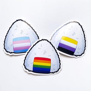 Può includere: Tre adesivi di palline di riso bianche con diverse bandiere pride. Il primo adesivo ha una bandiera pride transgender, il secondo ha una bandiera pride non binaria e il terzo ha una bandiera pride arcobaleno.
