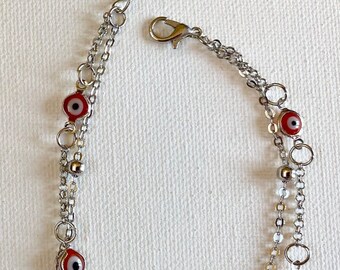 Puede incluir: Pulsera de cadena plateada con múltiples amuletos de ojo turco rojos y blancos. La pulsera tiene un diseño de doble hebra con pequeñas cuentas plateadas y eslabones circulares. Un cierre de mosquetón asegura la pulsera.