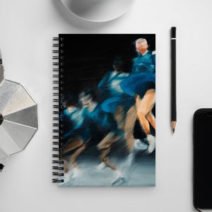 Könnte beinhalten: Spiralgebundenes Notizbuch mit einem abstrakten Gemälde von Tänzern in Bewegung, in Blau- und Weißtönen vor schwarzem Hintergrund. Das Notizbuch liegt auf einer weißen Oberfläche, zusammen mit einer Kaffeetasse, einem Bleistift, einem Telefon und einer silbernen Kaffeemaschine.