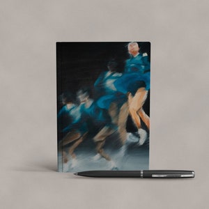 Könnte beinhalten: Ein schwarzes Notizbuch mit einem verschwommenen Bild von Eiskunstläufern in blauen Uniformen auf dem Cover. Ein schwarzer Stift liegt vor dem Notizbuch.