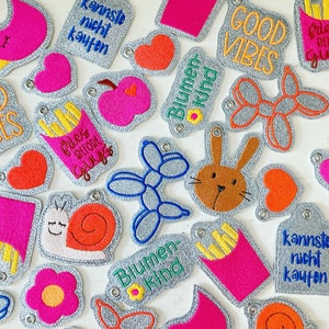 Può includere: Una collezione di toppe ricamate colorate con vari disegni, tra cui un cuore, una lumaca, un coniglio, un animale a palloncino, un fiore, una porzione di patatine fritte e testo che dice "Good Vibes", "Blumen Kind", "kannste nicht kaufen" e "Fles Before Guys".