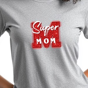 May include: A light gray t-shirt featuring a red glitter design that says "Super Mom" in white script. The "M" is a large, red glitter letter. The shirt is made of a soft material.