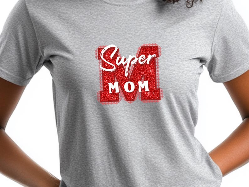 May include: A light gray t-shirt featuring a red glitter design that says "Super Mom" in white script. The "M" is a large, red glitter letter. The shirt is made of a soft material.