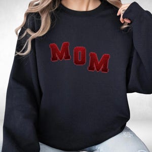 May include: Navy blue sweatshirt with the word "MOM" in red, block-letter style. The letters have a textured, slightly raised appearance. The sweatshirt is a casual, comfortable style.