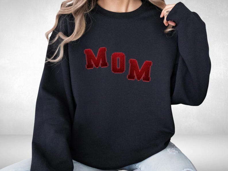 May include: Navy blue sweatshirt with the word "MOM" in red, block-letter style. The letters have a textured, slightly raised appearance. The sweatshirt is a casual, comfortable style.