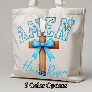 Op de afbeelding: Crèmekleurige draagtas met het woord "AMEN" in blauw omlijnde letters, gevuld met bloemmotieven. Een houten kruis met een lichtblauwe strik is gecentreerd onder het woord. De zin "He has Risen" is in lichtblauw schrift onder het kruis geschreven.