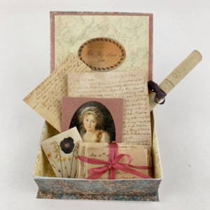 May include: A vintage-style box with a lid that opens to reveal a collection of antique-looking papers, a rolled-up scroll, and a framed portrait of a woman. The box is decorated with a floral pattern and the lid has a label that reads "To My Love, 1799".
