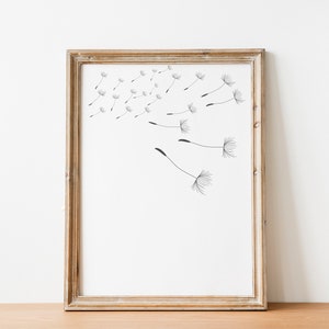 May include: A framed print of a black and white illustration of dandelion seeds blowing in the wind. The frame is a light brown wood.
