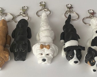 May include: A collection of five dog-shaped keychains. The keychains feature various breeds, including a brown and white dog, a black dog, a white dog with a bow, a black and white dog, and a black, white, and brown dog. Each keychain has a silver clasp.