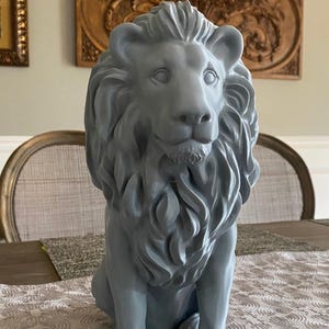 May include: A gray lion statue with a detailed mane and facial features. The sculpture is seated on a table with a patterned runner. Decorative wall art is visible in the background.
