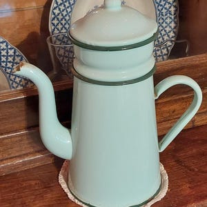 May include: A light blue enamel coffee pot with a lid, curved spout, and dark green trim. The pot has a handle and rests on a white, crocheted doily. The background features a wooden surface and decorative plates.