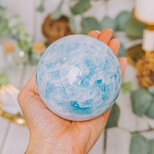 May include: A light blue aquamarine crystal sphere, with a smooth, polished surface, is held in a hand. The sphere is about the size of a baseball.