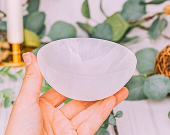 May include: A small, white, oval-shaped bowl with a smooth surface. The bowl is held in a hand, with a blurred background of greenery and a candle holder. The bowl is likely made of a stone material.
