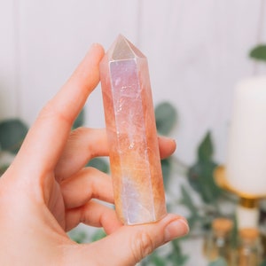 May include: A pink and orange crystal point, also known as a crystal wand, is held in a person's hand. The crystal is smooth and polished, with a pointed tip. The background is blurred and features greenery and candles.