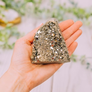 May include: A large, golden, pyramid-shaped pyrite crystal with a rough, textured surface. The crystal is being held in a person's hand.