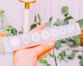 May include: A white selenite wand with seven etched chakra symbols. The wand is held in a hand against a blurred background of greenery and candles.
