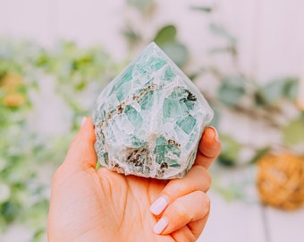 May include: A green fluorite crystal point with a pointed top and a rough, textured surface. The crystal is held in a hand.