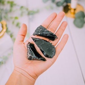 Puede incluir: Tres piezas de obsidiana negra, una roca volc&aacute;nica oscura y vidriosa, se sostienen en una mano. Las piezas tienen una forma irregular y bordes afilados.