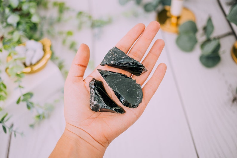 Puede incluir: Tres piezas de obsidiana negra, una roca volc&aacute;nica oscura y vidriosa, se sostienen en una mano. Las piezas tienen una forma irregular y bordes afilados.