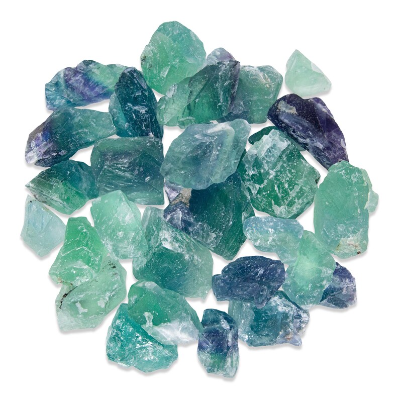 May include: A collection of raw fluorite crystals in shades of green, blue, and purple. The natural, rough-cut stones vary in size and shape, with a translucent quality. The crystals are scattered against a white background.