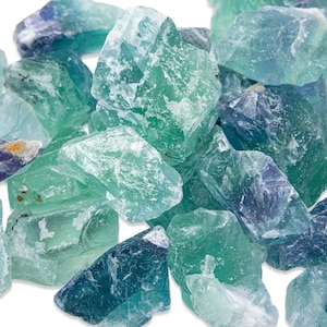 May include: A collection of raw fluorite crystals in shades of green, blue, and purple. The rough, natural stones have a translucent quality, with varying depths of color and visible internal structures. The crystals are piled together against a white background.